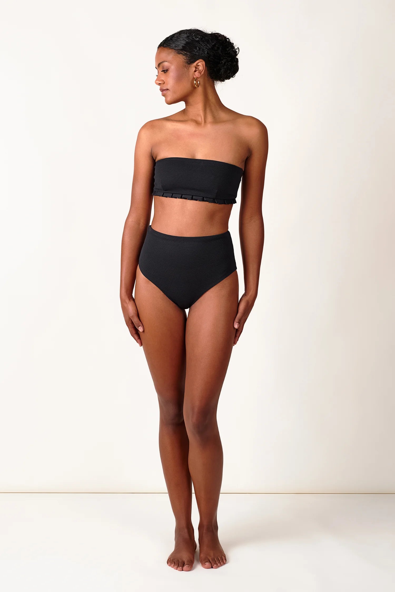 Frau trägt schwarzen MYMARINI Bandeau-Bikinitop und High-Waist-Bikinihose vor hellem Hintergrund.#color_black