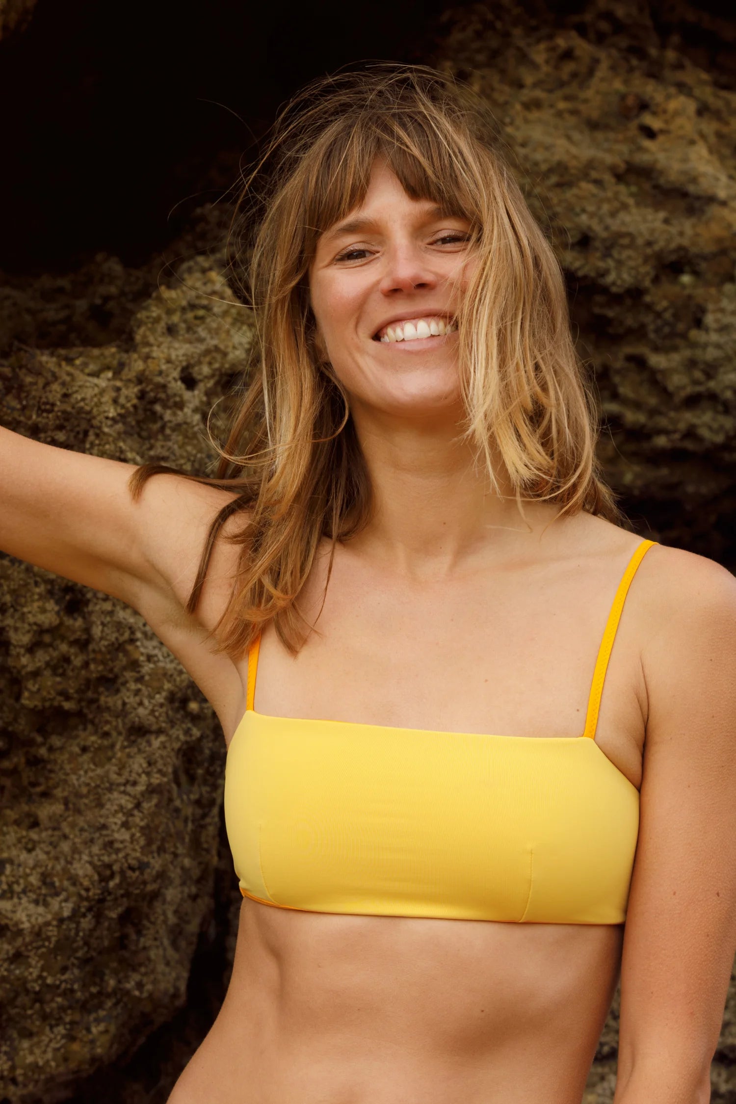 Frau am Strand trägt ein gelbes MYMARINI Bikinitop mit Spaghettiträgern. Sie lächelt vor Felsen.#color_buttermilk-soleil