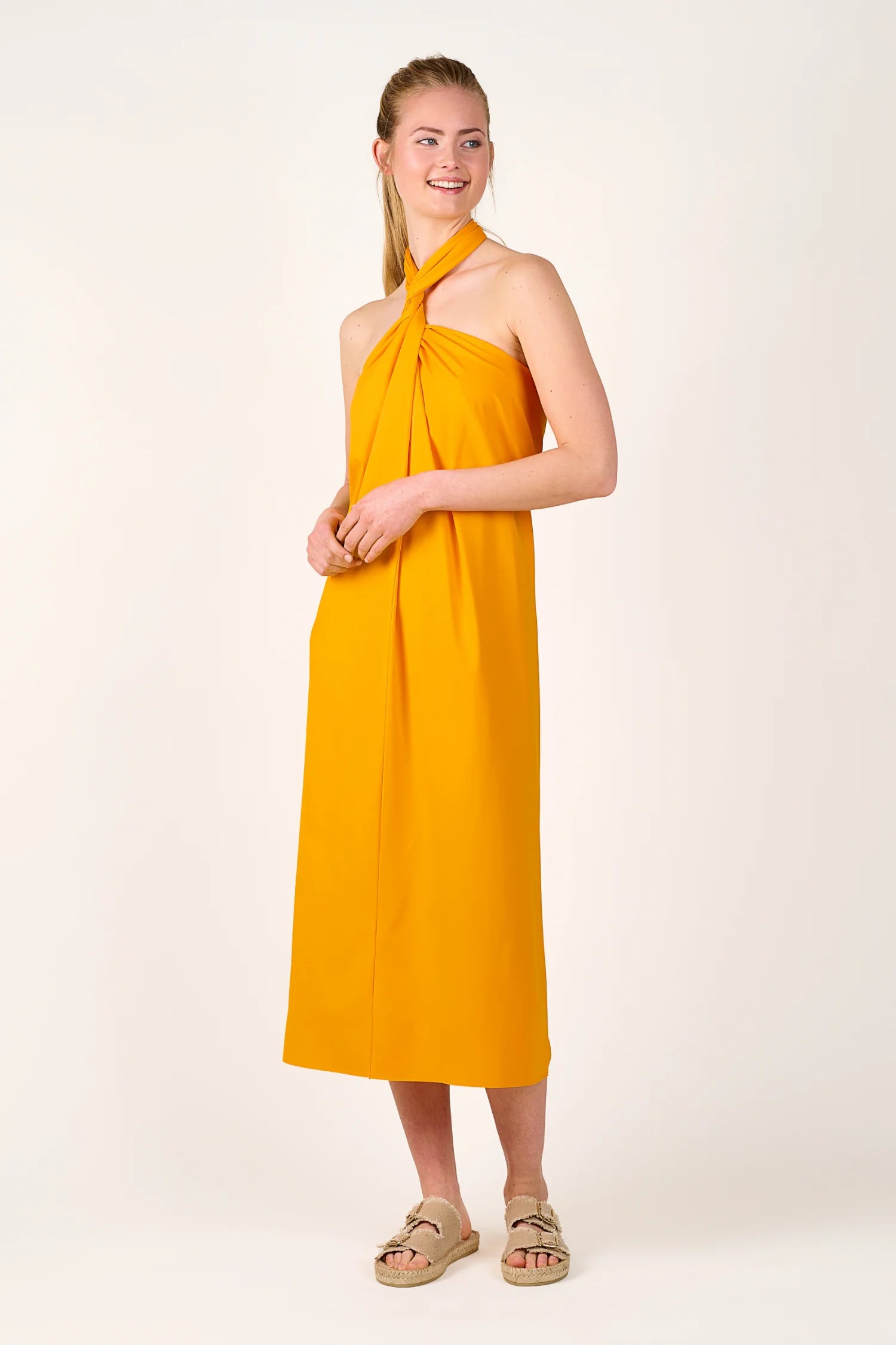 Person trägt ein gelbes MYMARINI-Kleid mit Neckholder-Schnitt, stehend vor neutralem Hintergrund.#color_buttermilk-soleil