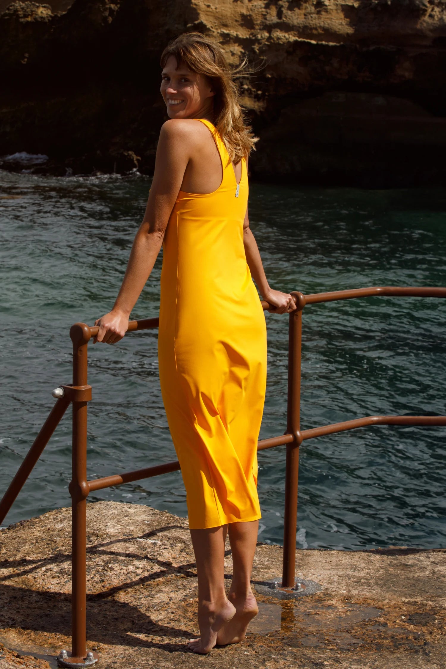 Frau in gelbem MYMARINI-Kleid mit breiten Trägern steht am Geländer vor dem Meer.#color_buttermilk-soleil