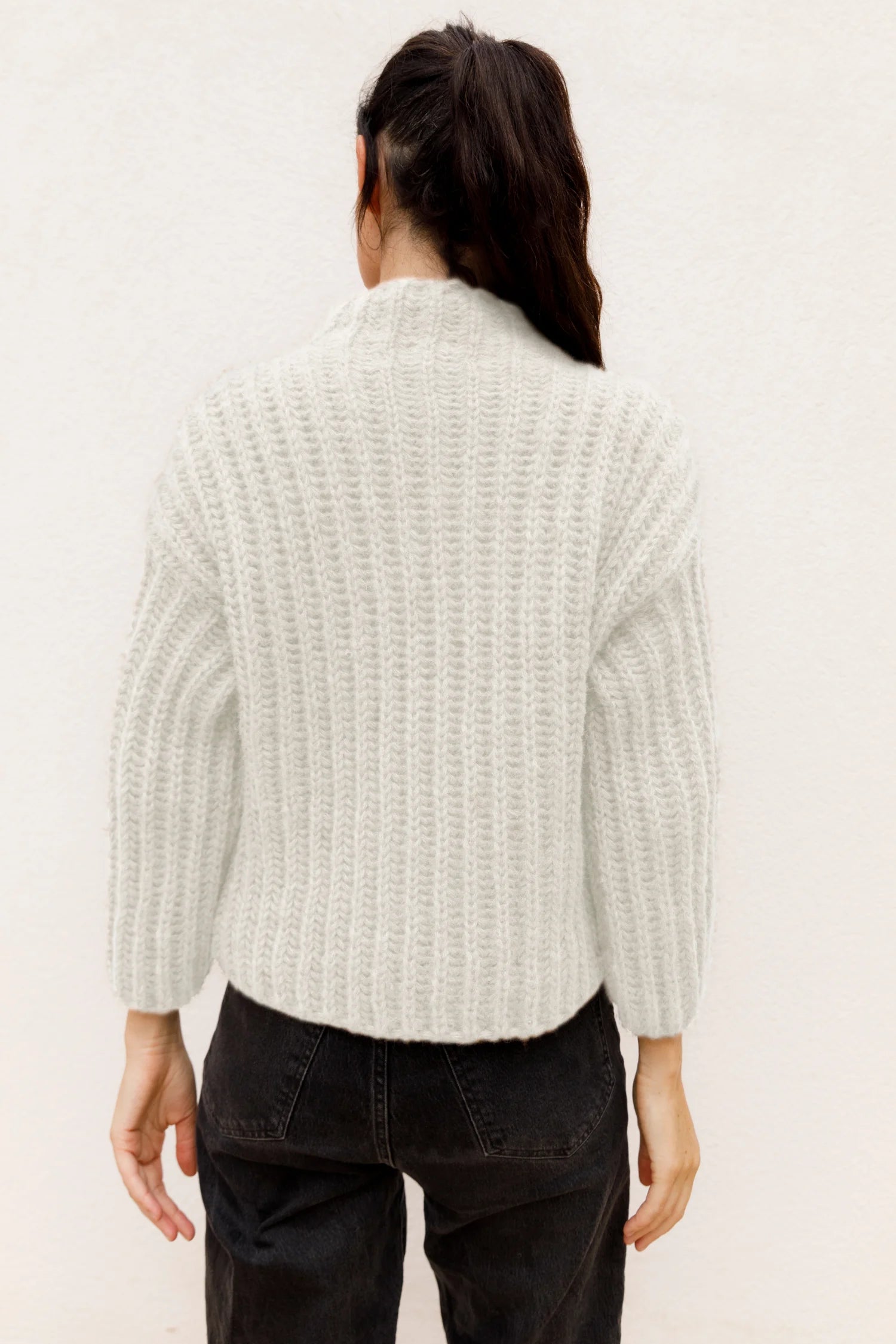 Person von hinten in weißem Strickpullover und schwarzer Hose vor hellem Hintergrund.#color_white-stone
