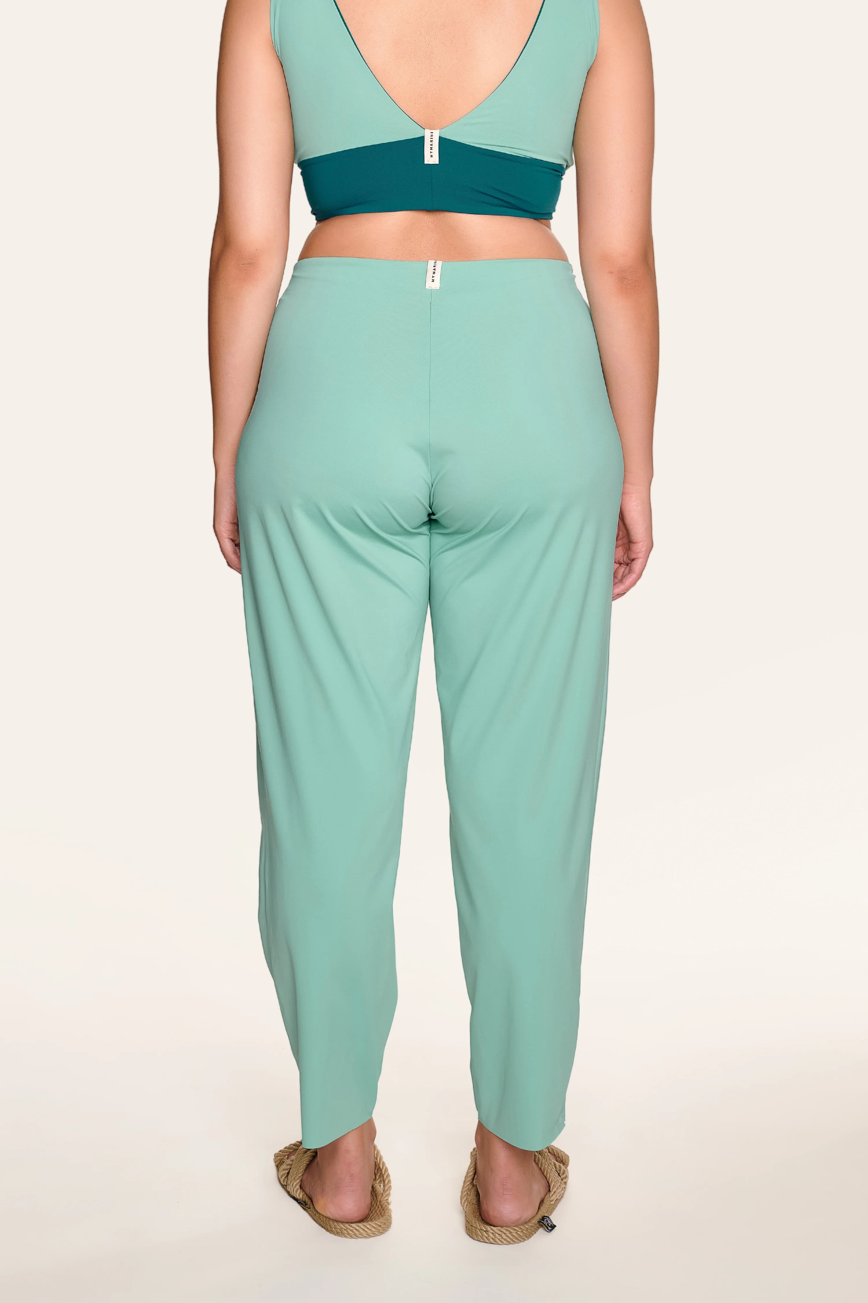 Rückansicht einer Person in MYMARINI-Hose und Bikinitop mit V-Ausschnitt, breiten Trägern und flachen Sandalen.#color_eucalyptus