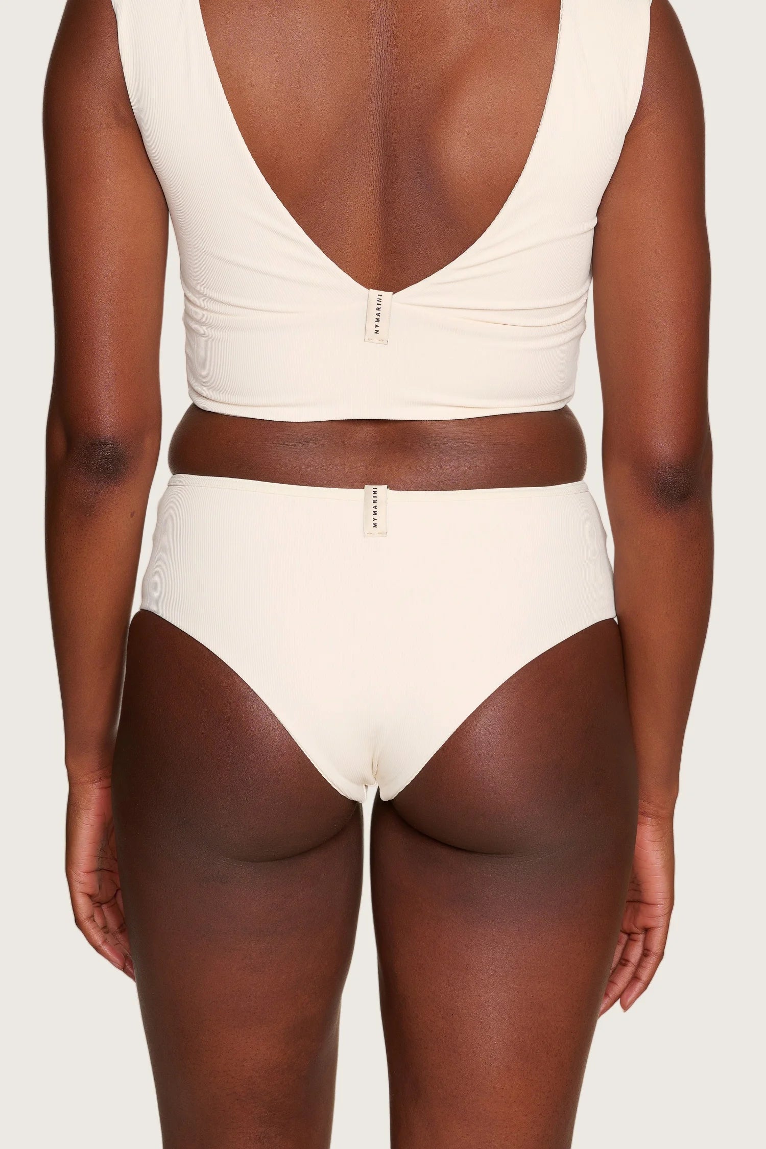 Rückansicht einer Person in cremefarbenem MYMARINI-Bikinitop mit tiefem Rückenausschnitt und passender Bikinihose, mid rise.#color_off-white