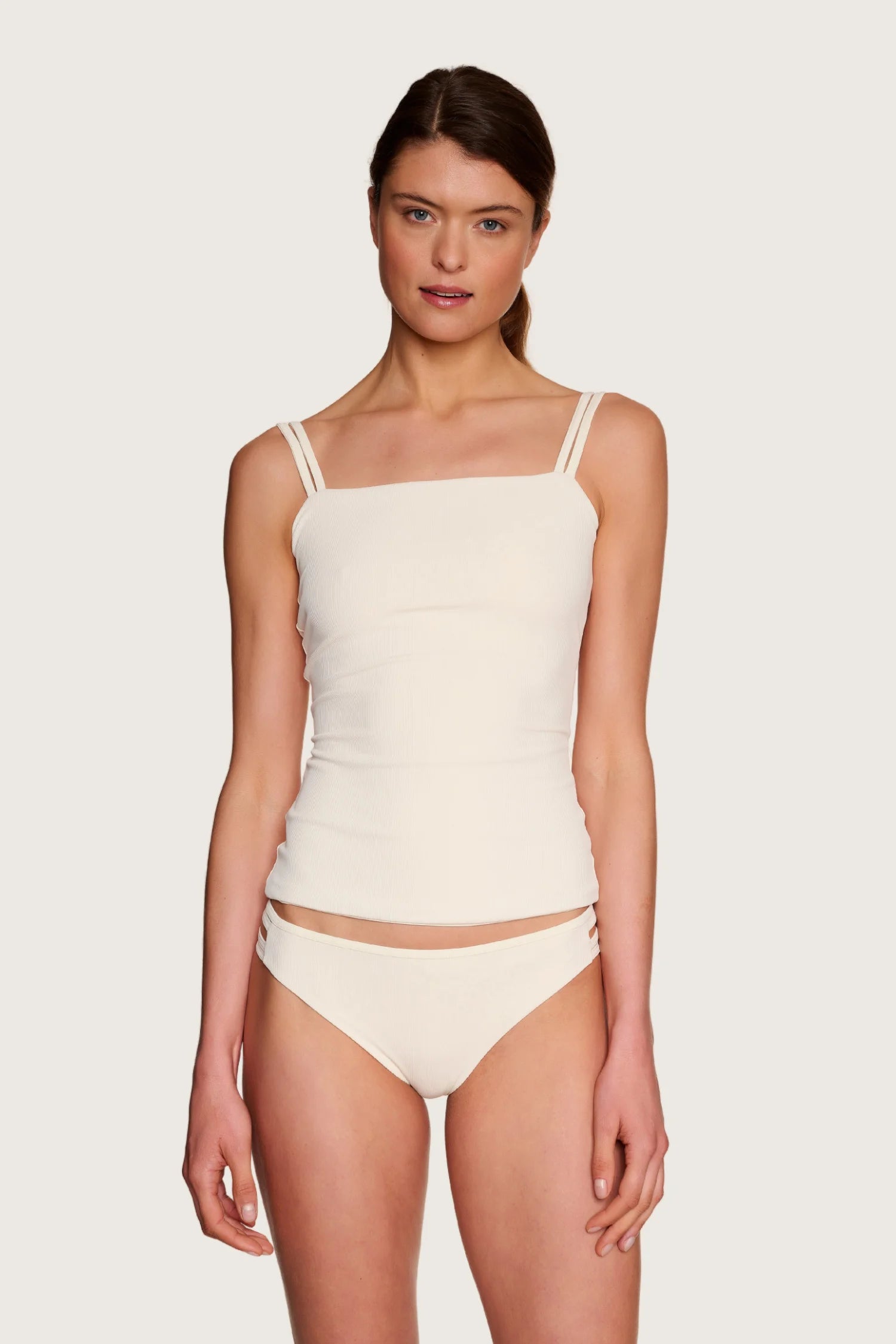 Frau trägt MYMARINI Bikinihose mit hohem Beinausschnitt und Top mit Spaghettiträgern in Cremeweiß vor neutralem Hintergrund.#color_off-white
