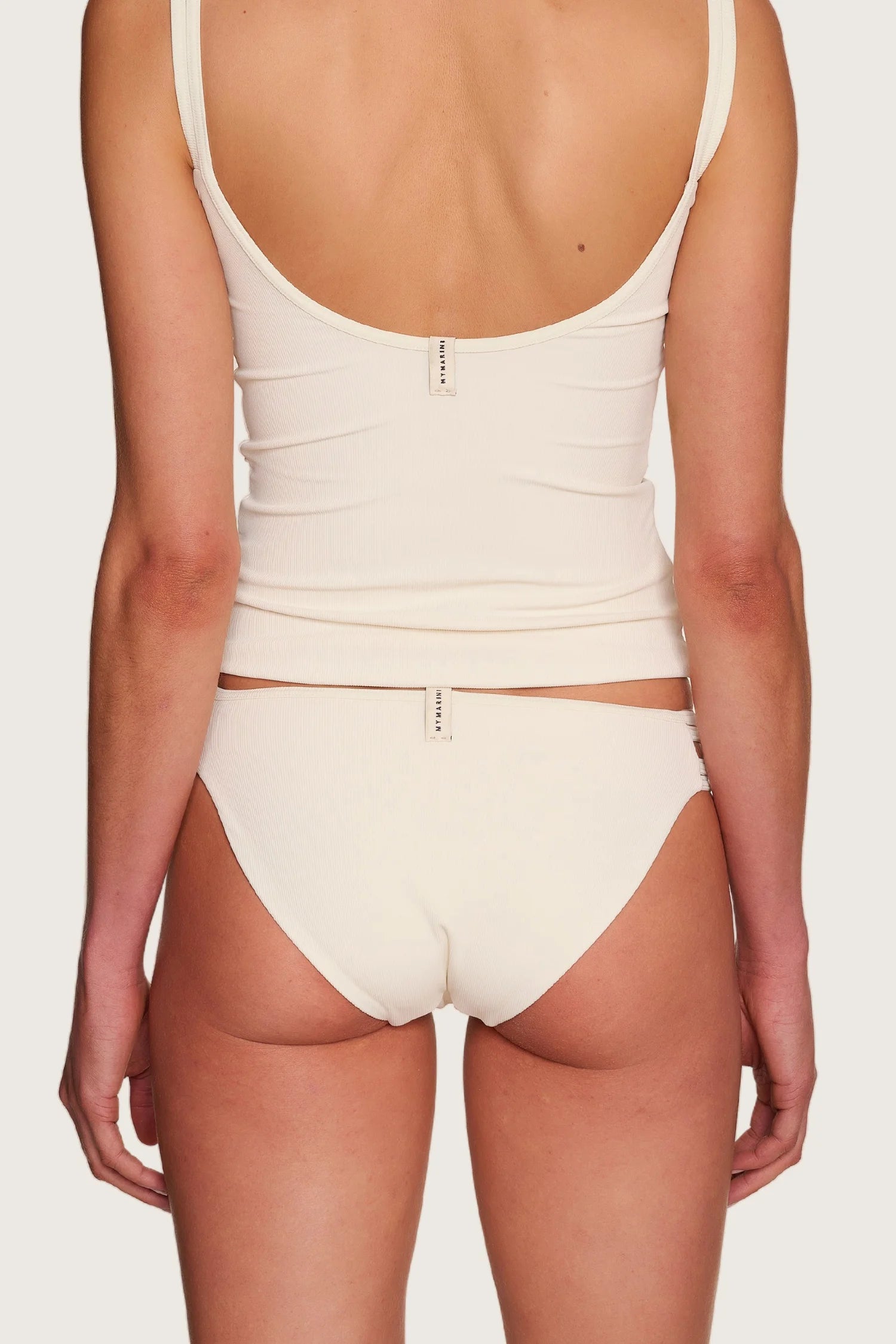 Rückansicht einer Person in cremefarbenem MYMARINI-Top mit tiefem Rückenausschnitt und passender Bikinihose, hoher Beinausschnitt.#color_off-white
