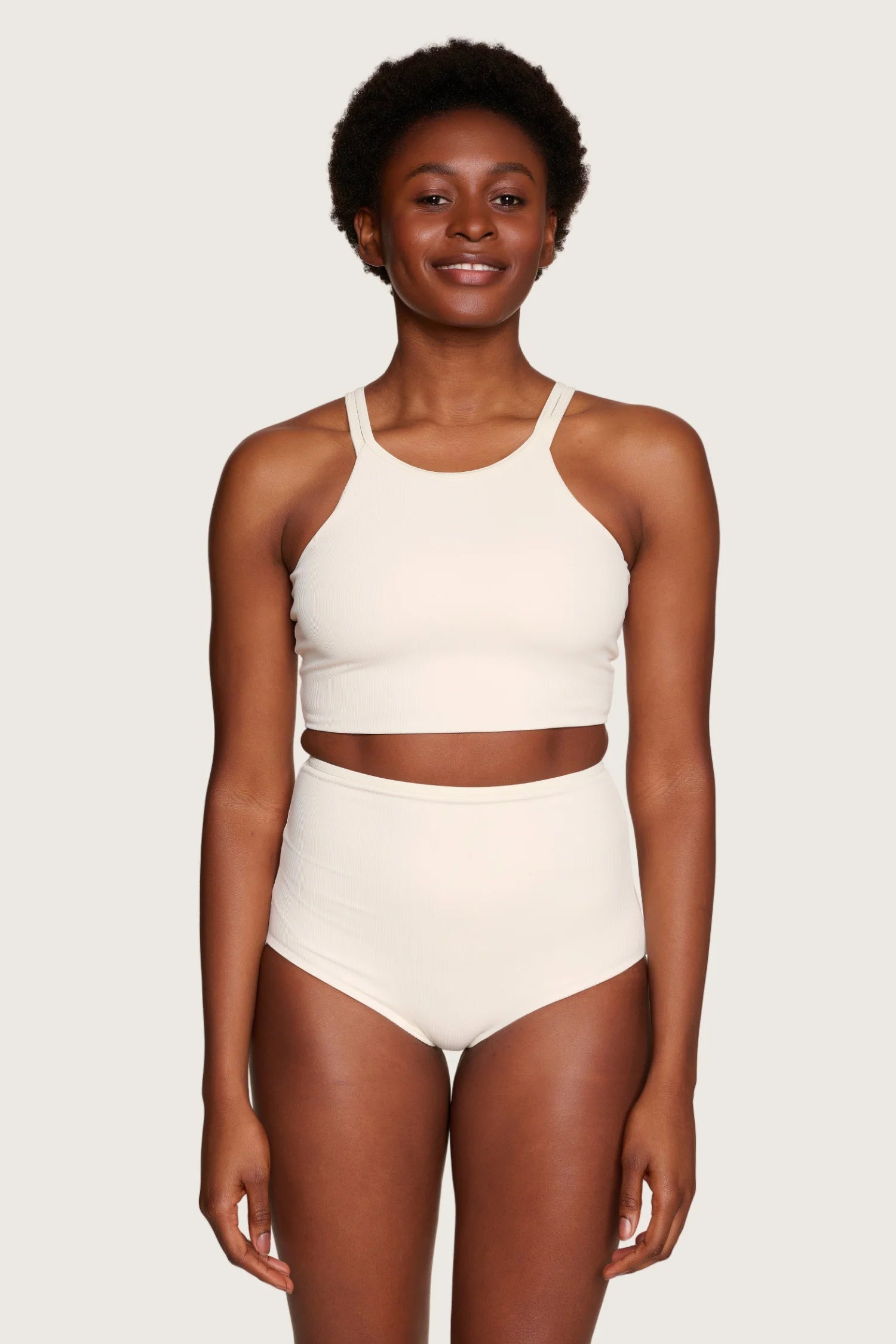 Frau trägt MYMARINI Bikini mit hochgeschlossenem Top und High-Waist-Hose in Creme vor neutralem Hintergrund.#color_off-white
